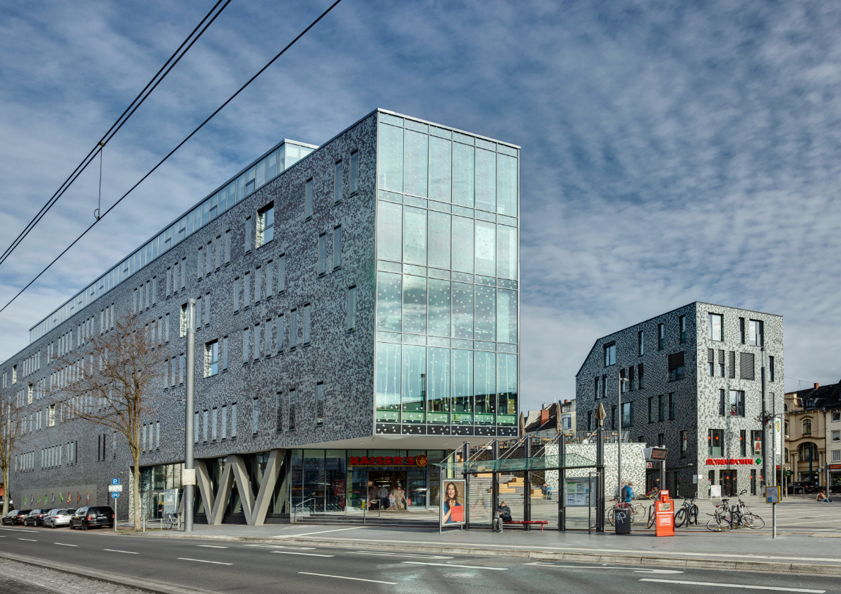 Mehrgeschossiges Gebäude mit markanter Glasfassade an einer Straßenecke im städtischen Kontext. Facharztzentrum mit einem Vollsortimenter mit prägnanter Keramikfassade und transparenter Fassadenlösung in Bonn-Beuel.