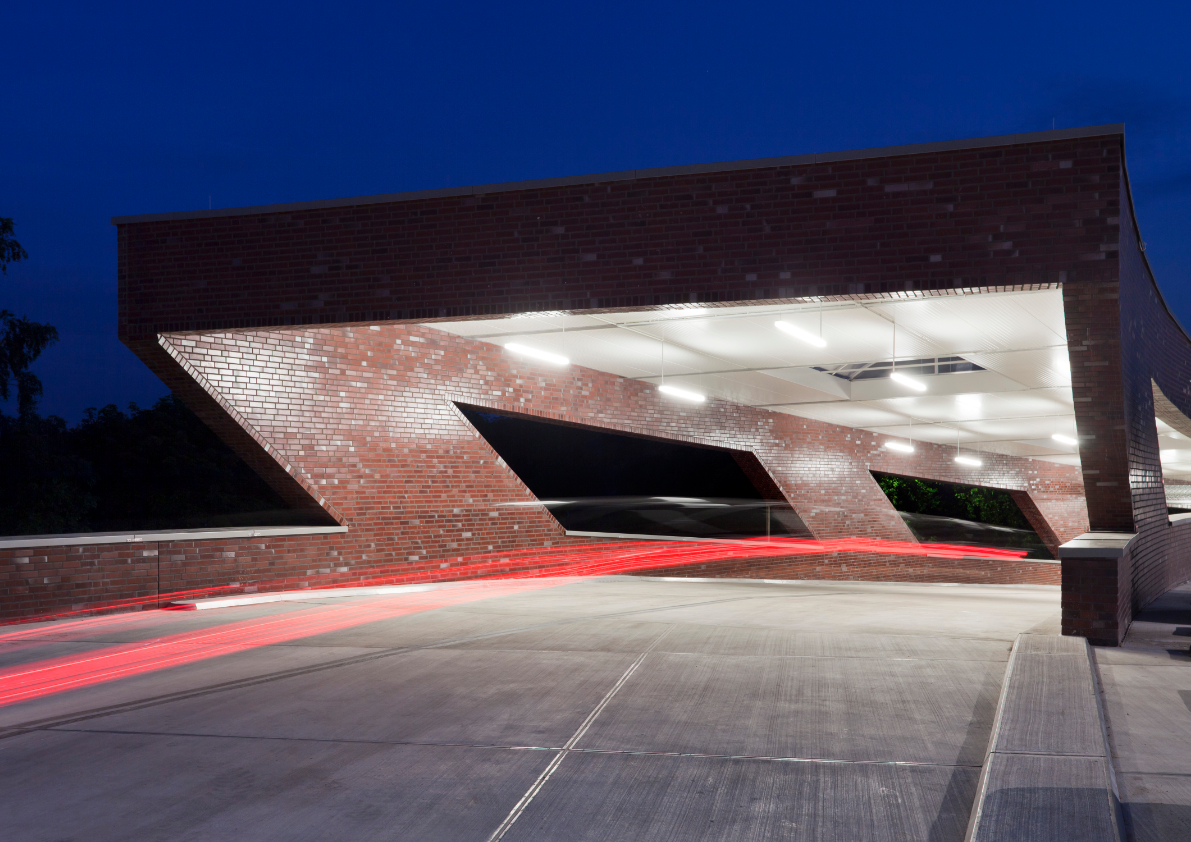 Backsteinbau mit geneigten Öffnungen und beleuchteter Durchfahrt bei Nacht, mit Lichtspuren von Fahrzeugen