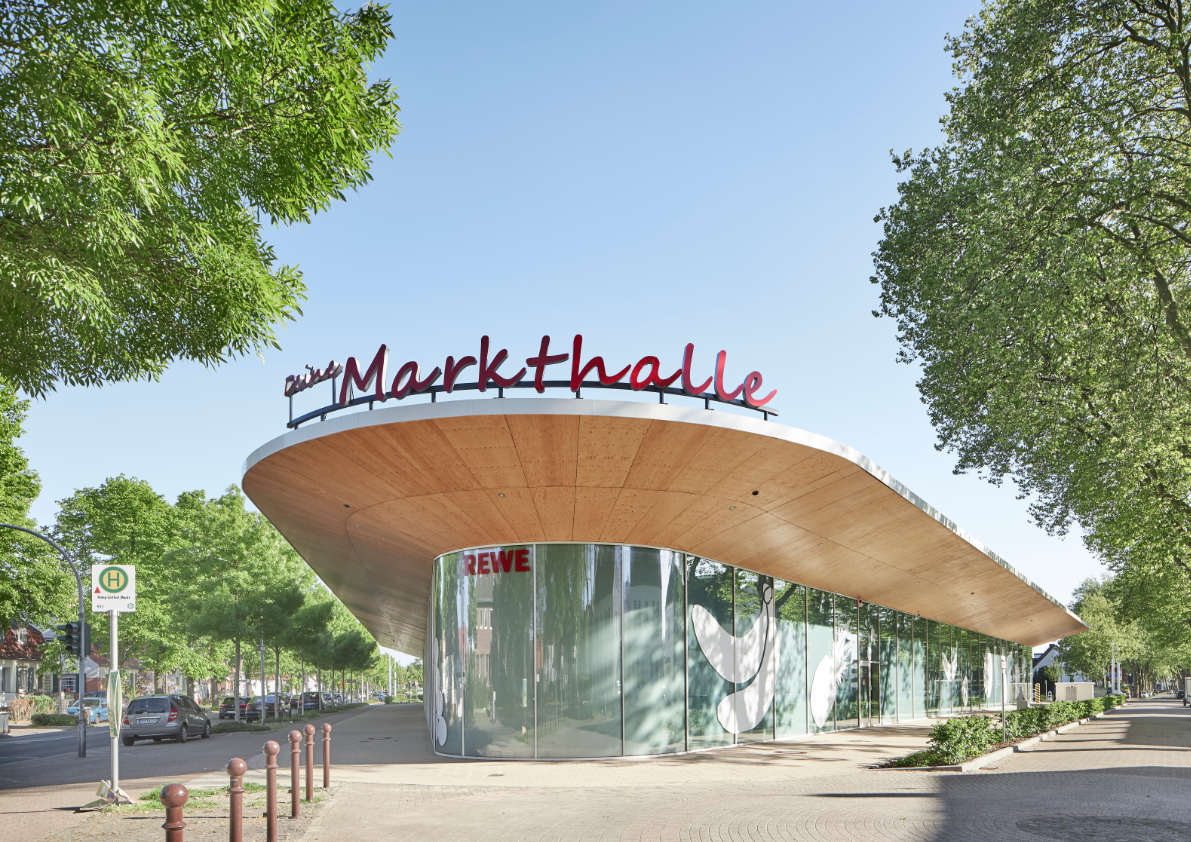 Supermarktgebäude REWE mit geschwungener Dachform auf dem ehemaligen Marktplatz in der Altsiedlung von Kamp-Lintfort mit markanter Grafik auf der transluzenten Fassade.