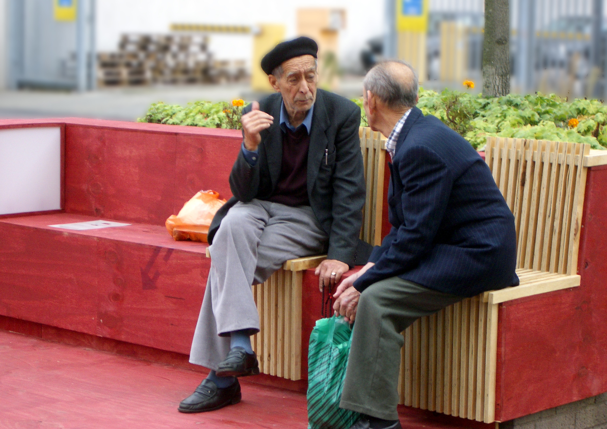 Zwei ältere Männer sitzen auf einer roten Sitzstruktur im öffentlichen Raum am Neptunplatz in Köln Ehrenfeld